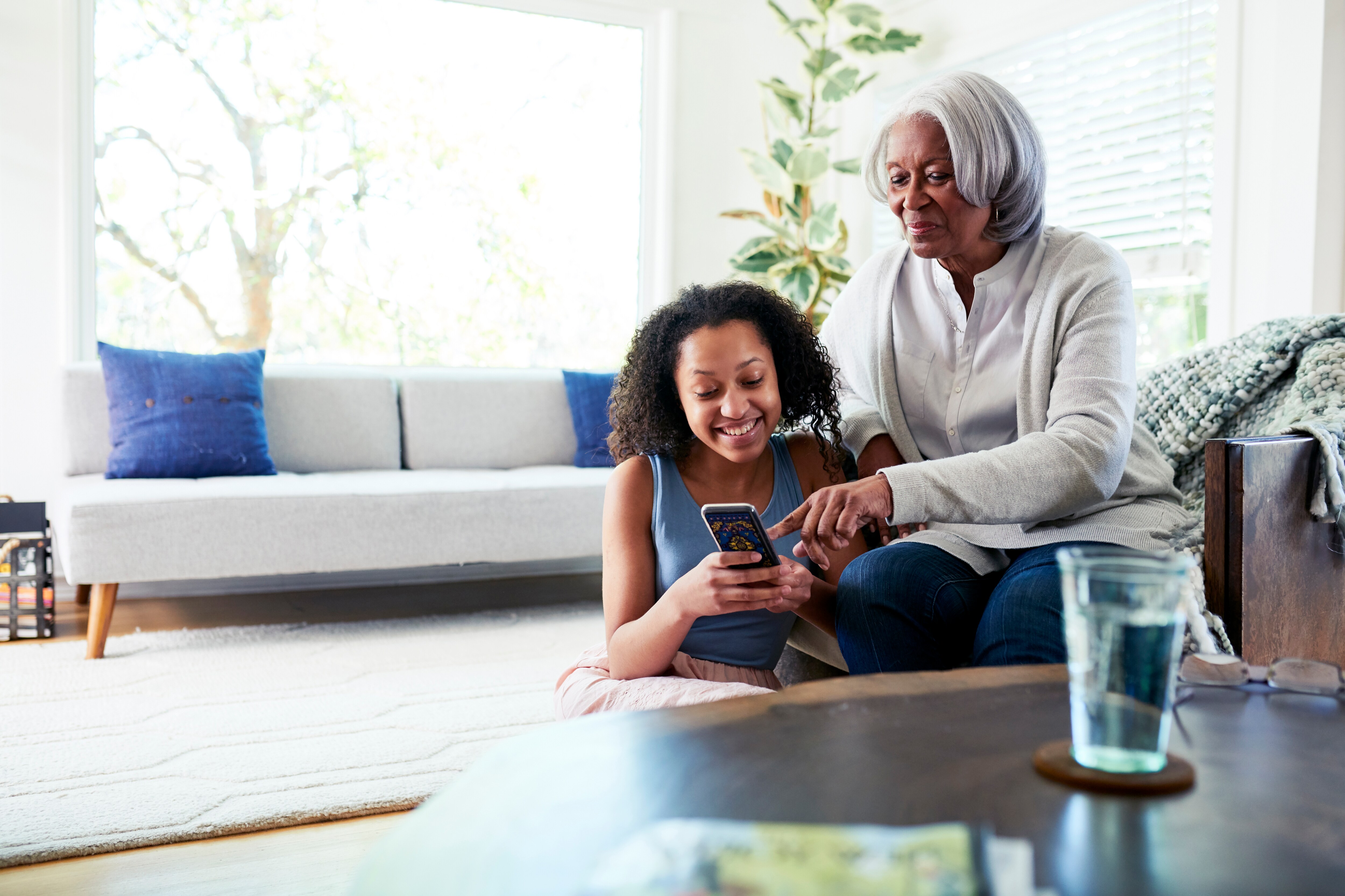 older individual seated on a couch next to a younger child who is sitting on the floor and both are looking at a smartphone together in a bright living room